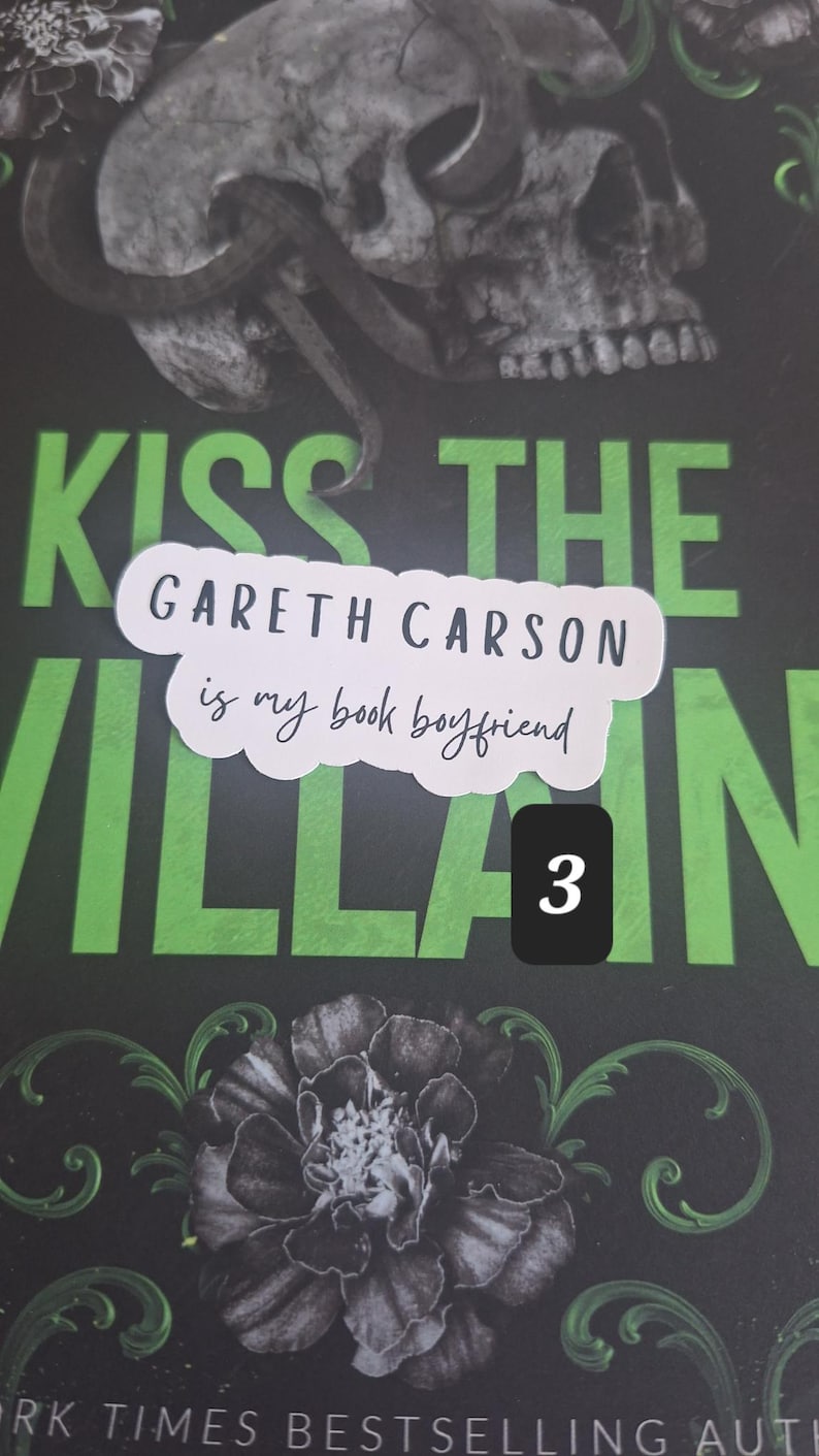May include: A book cover with the title "Kiss the Villain" in green lettering. A sticker on the cover reads "Gareth Carson is my book boyfriend." The cover features a skull, snake, and floral design.