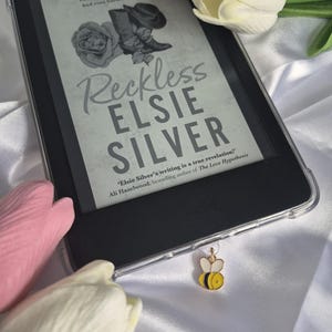 May include: A black e-reader displaying the book cover of "Reckless" by Elsie Silver. The cover art features a rose, boots, and a hat. A bee charm hangs from the e-reader. Pink and white tulips are in the background.