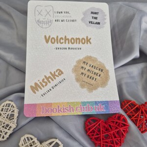 May include: A white rectangular card with text and stickers, including the title "Volchonok" and author's name. The card has a rainbow-colored stripe at the bottom with the text "bookish club uk". Decorative red and white hearts are in the foreground.