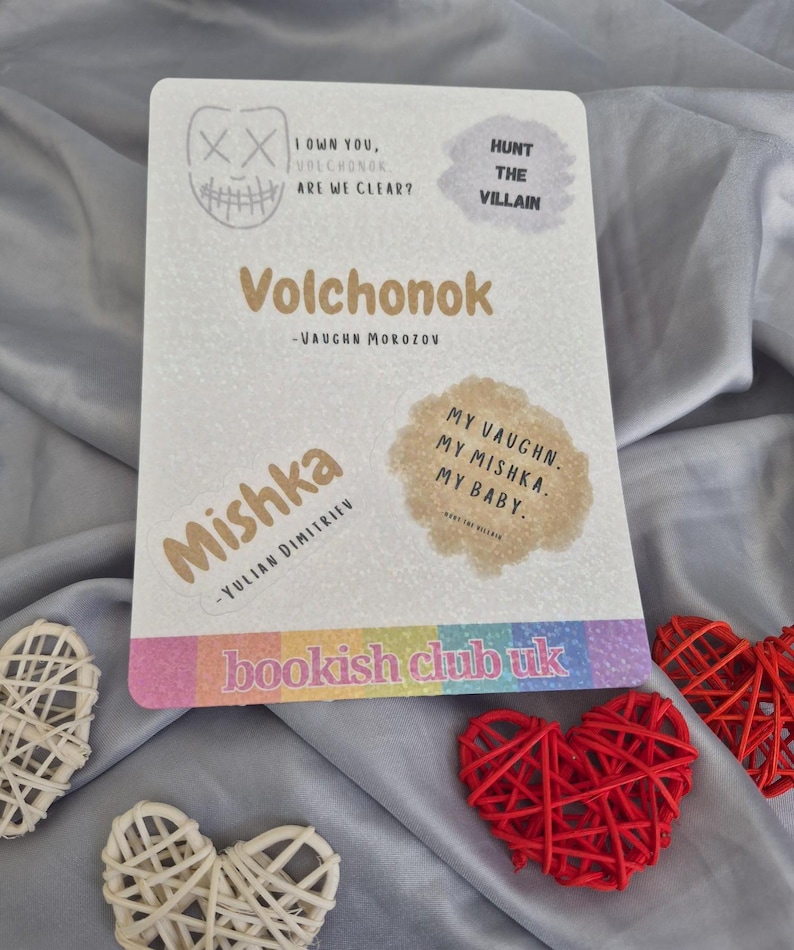May include: A white rectangular card with text and stickers, including the title "Volchonok" and author's name. The card has a rainbow-colored stripe at the bottom with the text "bookish club uk". Decorative red and white hearts are in the foreground.