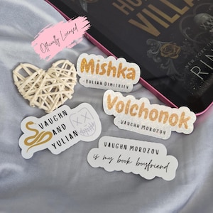May include: A collection of stickers with text and a woven heart. The stickers have gold and white text, including names and phrases like "Mishka" and "Vaughn Morozov is my book boyfriend." An e-reader and book are in the background.