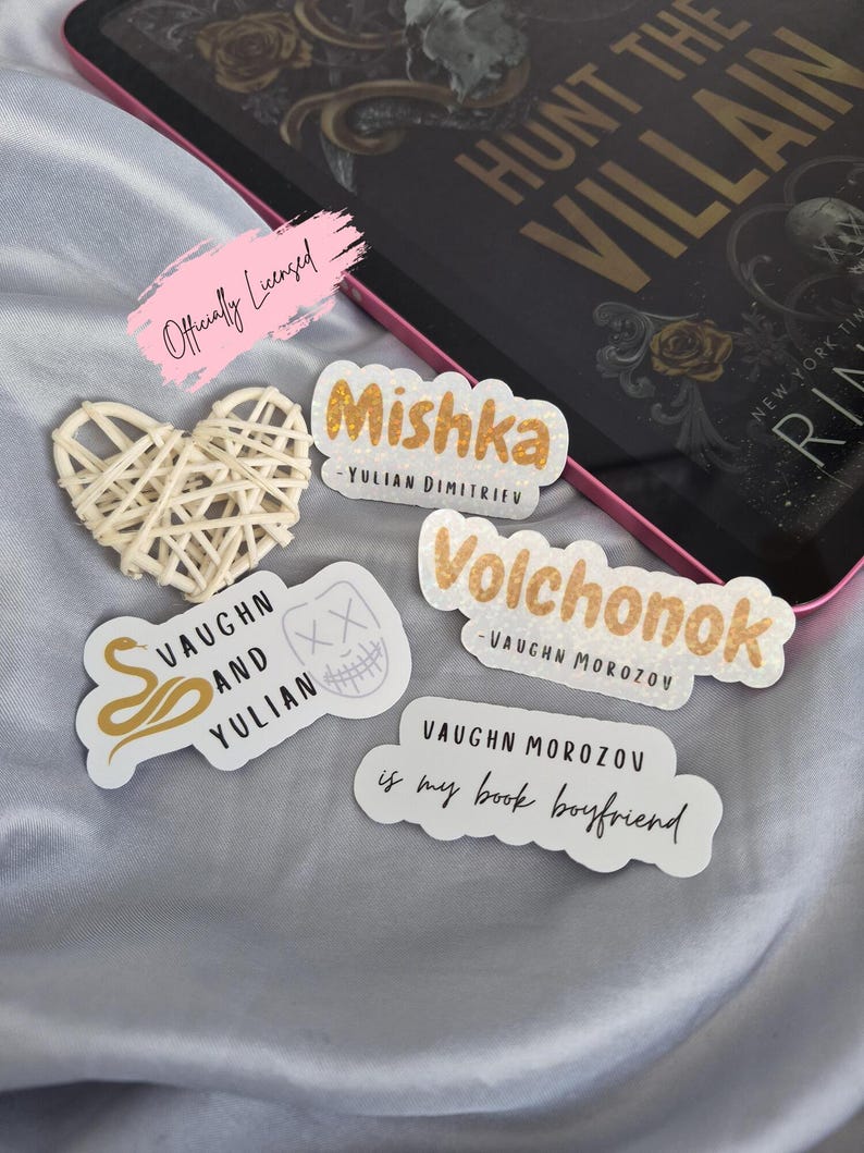 May include: A collection of stickers with text and a woven heart. The stickers have gold and white text, including names and phrases like "Mishka" and "Vaughn Morozov is my book boyfriend." An e-reader and book are in the background.