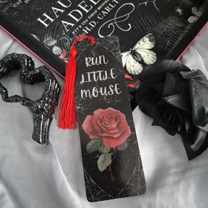 May include: A bookmark with a red tassel, featuring a red rose illustration and the text "Run Little Mouse" on a black background. The bookmark is next to a book titled "Haunting Adeline" and a black decorative heart.