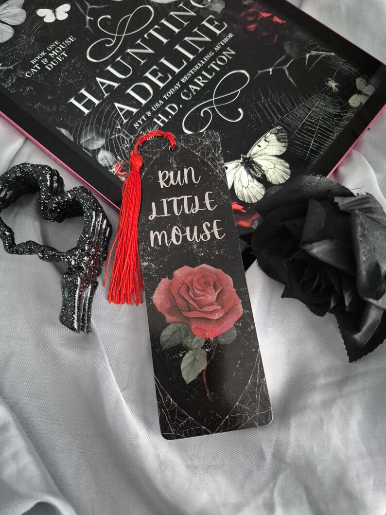 May include: A bookmark with a red tassel, featuring a red rose illustration and the text "Run Little Mouse" on a black background. The bookmark is next to a book titled "Haunting Adeline" and a black decorative heart.