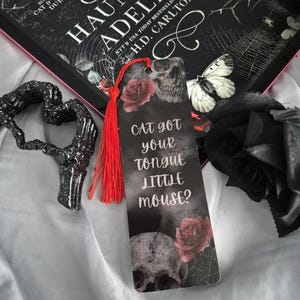May include: A bookmark with a skull and rose design, the text "Cat got your tongue little mouse?" and a red tassel. The bookmark is next to a book titled "Haunting Adeline" and a black heart-shaped decoration.
