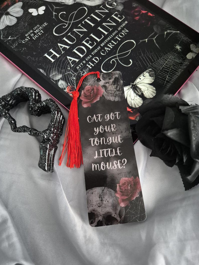 May include: A bookmark with a skull and rose design, the text "Cat got your tongue little mouse?" and a red tassel. The bookmark is next to a book titled "Haunting Adeline" and a black heart-shaped decoration.