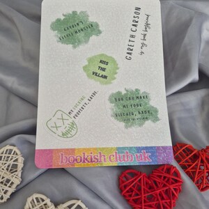 May include: A rectangular white sticker with green text bubbles and the text "bookish club uk". The sticker has phrases like "Kayden's Little Monster" and "Kiss the Villain". Decorative red and white woven hearts are in the foreground.