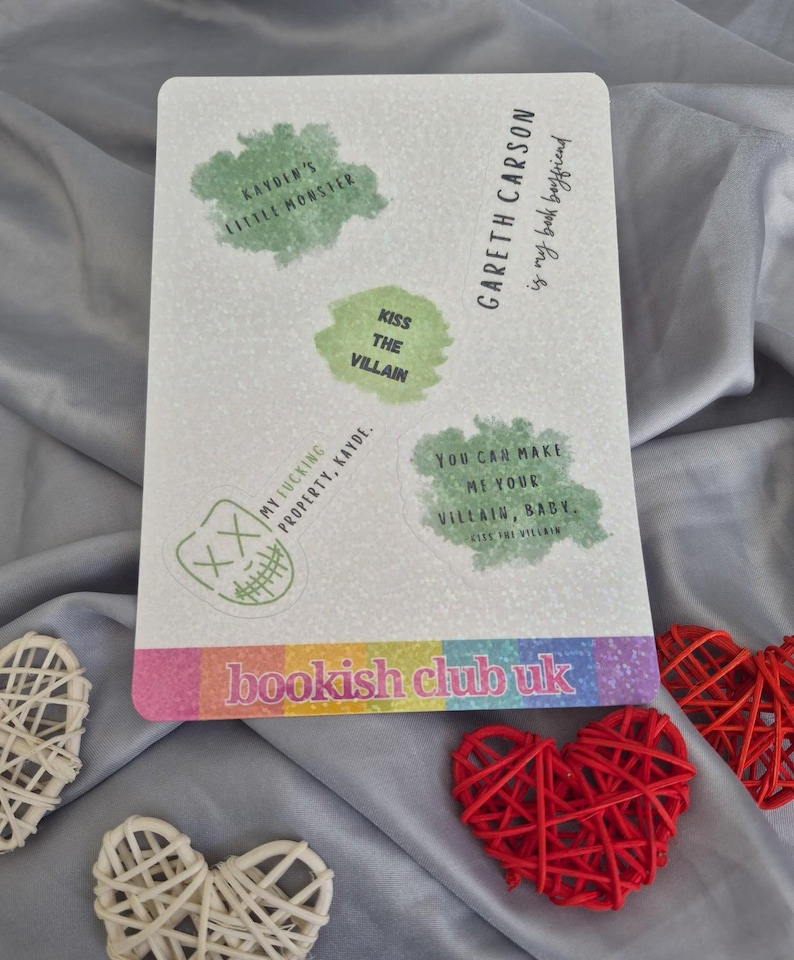 May include: A rectangular white sticker with green text bubbles and the text "bookish club uk". The sticker has phrases like "Kayden's Little Monster" and "Kiss the Villain". Decorative red and white woven hearts are in the foreground.