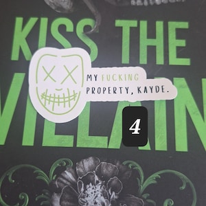 May include: A book cover with the title "Kiss the Villain" in large green letters. A skull and snake graphic is at the top. A sticker with a mask design and the text "My Fucking Property, Kayde" is on the cover. A black square with the number 4 is also present.