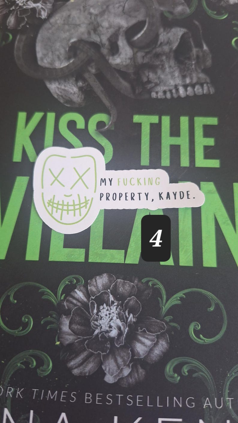 May include: A book cover with the title "Kiss the Villain" in large green letters. A skull and snake graphic is at the top. A sticker with a mask design and the text "My Fucking Property, Kayde" is on the cover. A black square with the number 4 is also present.
