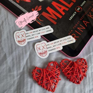 May include: The image features two red woven heart decorations, two stickers with a smiling face design and the text "YOU CAN BREAK MY LEGS AND ARMS, BUT I CAN STILL CRAWL TOWARDS HER.", and a book titled "Legacy of Gods".