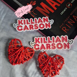 May include: Red woven heart-shaped decorations and stickers with the text "Killian Carson" in red and white, with heart accents. A book with the title "Malice" is visible in the background.