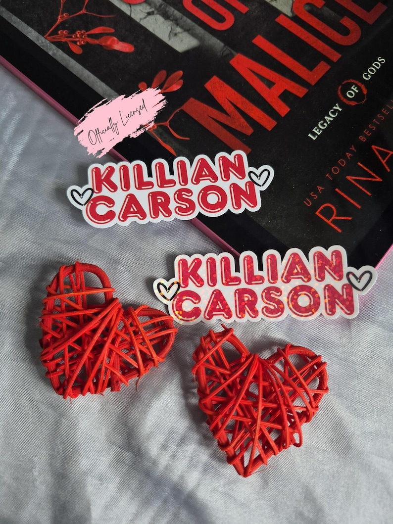 May include: Red woven heart-shaped decorations and stickers with the text "Killian Carson" in red and white, with heart accents. A book with the title "Malice" is visible in the background.