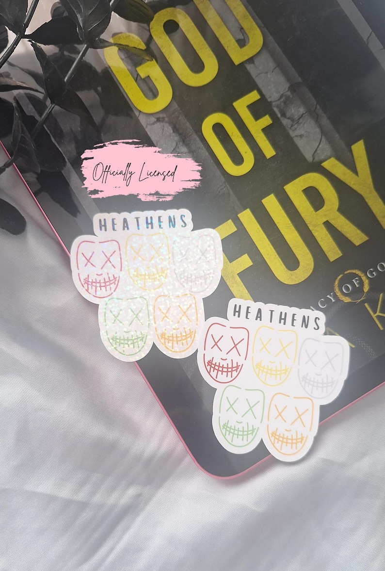 May include: Two stickers with a design of colorful masks with an 'XX' on the eyes and a stitched smile. The word 'HEATENS' is printed above the masks. The stickers are on top of a book with the title 'GOD OF FURY'.