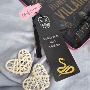 May include: A black bookmark with a white graphic of a smiling face and the words "Volchonok and Mishka" and a gold snake design. Two woven heart decorations are next to the bookmark. The bookmark is next to a book with the title "Hunt the Villain".