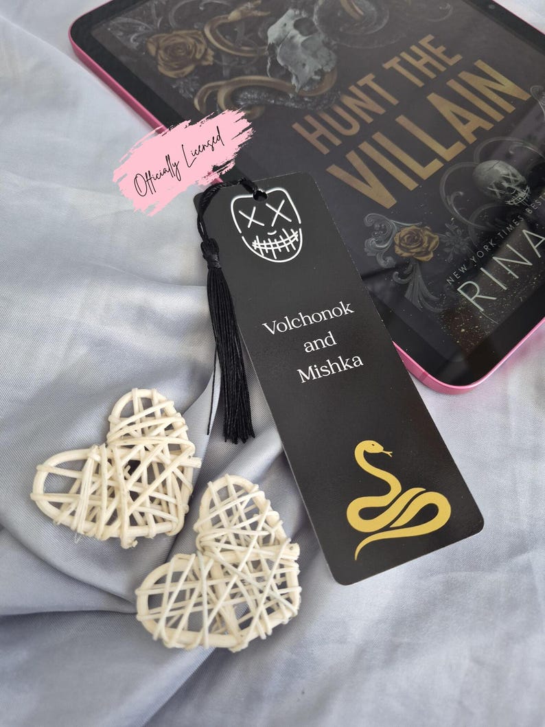 May include: A black bookmark with a white graphic of a smiling face and the words "Volchonok and Mishka" and a gold snake design. Two woven heart decorations are next to the bookmark. The bookmark is next to a book with the title "Hunt the Villain".