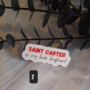 May include: White sticker with red text that says "SAINT CARTER is my book boyfriend". The sticker is on a gray surface with a black branch in the background. The number "1" is written in red on a white square.