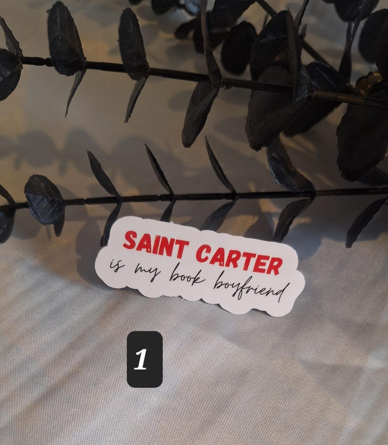 May include: White sticker with red text that says "SAINT CARTER is my book boyfriend". The sticker is on a gray surface with a black branch in the background. The number "1" is written in red on a white square.
