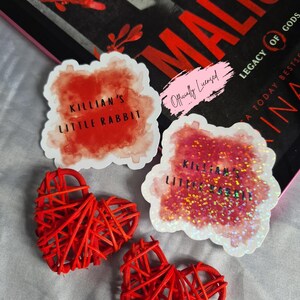 May include: An assortment of items including two red woven heart decorations, and two stickers with the text "KILLIAN'S LITTLE RABBIT". One sticker has glitter accents. A book with the title "OF MALICE" is in the background.