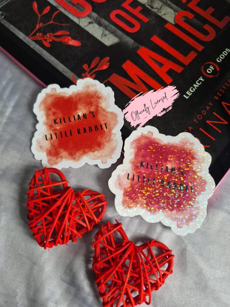 May include: An assortment of items including two red woven heart decorations, and two stickers with the text "KILLIAN'S LITTLE RABBIT". One sticker has glitter accents. A book with the title "OF MALICE" is in the background.