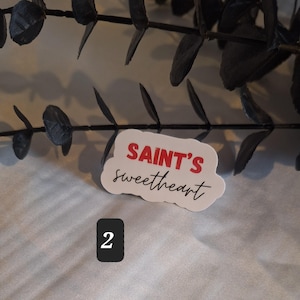 May include: White sticker with red text that says "SAINT'S sweetheart" in a cloud-like shape. The sticker is on a white surface with a number "2" written in black below it.