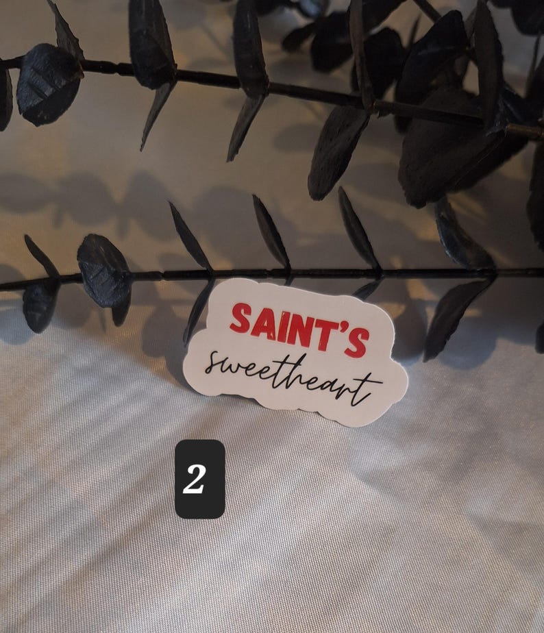 May include: White sticker with red text that says "SAINT'S sweetheart" in a cloud-like shape. The sticker is on a white surface with a number "2" written in black below it.