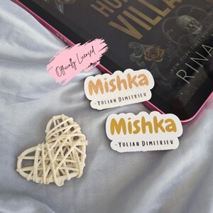 May include: A book titled "Hunt the Villain" with gold lettering and floral accents, a pink-edged tablet, two "Mishka" stickers, and a woven heart on a light blue surface. The stickers have the author's name.