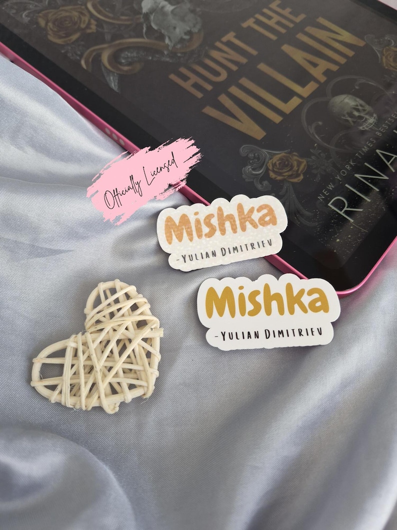 May include: A book titled "Hunt the Villain" with gold lettering and floral accents, a pink-edged tablet, two "Mishka" stickers, and a woven heart on a light blue surface. The stickers have the author's name.