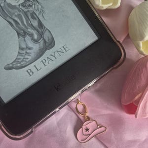 May include: A black e-reader in a clear protective case featuring a screen display of cowboy boots with roses and the text B L PAYNE. A gold-toned metal charm shaped like a pink cowboy hat with a small star detail is attached to the bottom edge.