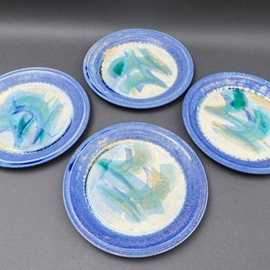 Op de afbeelding: Set van vier keramische borden met een blauwe rand en een wit centrum. De borden hebben een uniek abstract ontwerp met blauwe, groene en gouden accenten.