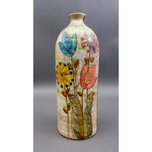 May include: A tall, ceramic vase with a cream-coloured background and colourful floral designs. The flowers are painted in shades of blue, yellow, red, and purple. The vase has a narrow neck and a slightly textured surface, adding to its handcrafted appearance.