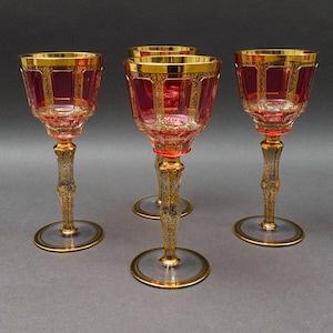 Puede incluir: Cuatro elegantes copas de vino con cuencos de color rojo rubí y bordes y tallos dorados ornamentados. Las copas tienen una base transparente con un borde dorado. Los tallos están intrincadamente detallados con un patrón dorado. Las copas están sobre un fondo gris.