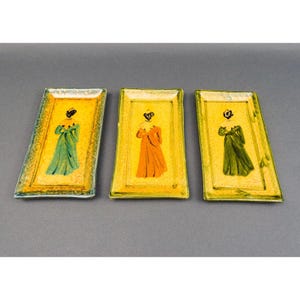 Puede incluir: Tres bandejas decorativas rectangulares de vidrio. Cada bandeja presenta un fondo amarillo con un patrón texturizado y una figura pintada con un vestido largo. Los vestidos son verdes y naranjas. Las bandejas tienen un borde transparente y texturizado.