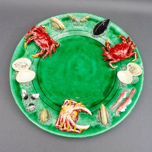 May include: A round, glossy green ceramic serving platter featuring three-dimensional relief sculptures of various seafood, including two bright red crabs, a smaller patterned crab, open clam shells, mussels, and shrimp arranged along the rim.