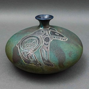 May include: A small, round, green and brown ceramic vase with a blue rim. The vase has a stylized animal design in black and white.