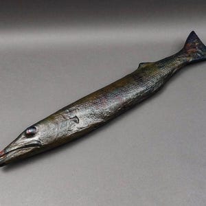May include: A long, dark green and brown fish sculpture with a detailed, textured surface. The fish has a long, pointed snout and a small, forked tail.