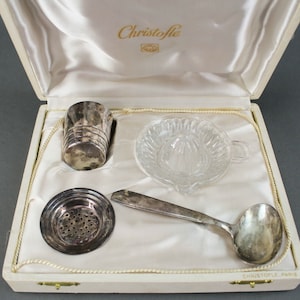 Puede incluir: Una caja blanca con un forro de satén que contiene un exprimidor de cítricos plateado, un colador plateado, una taza medidora plateada y una cuchara plateada. La caja está etiquetada con "Christofle" en la parte superior.