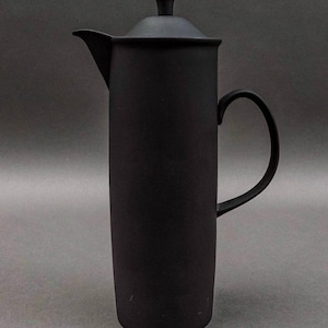 Op de afbeelding: Een matzwarte cafetière met een lange, cilindrische body, een deksel en een gebogen handvat. De tuit bevindt zich aan de linkerkant. De cafetière is ontworpen voor het zetten van koffie. De achtergrond is effen grijs.