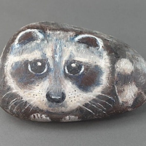 May include: Hand-painted rock art featuring a raccoon with black eyes, a white nose, and a gray and brown face. The raccoon is painted on a gray rock.