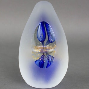 Op de afbeelding: Een mat wit glazen sculptuur met een blauw en gouden glazen interieur. Het interieur heeft een draaiend blauw ontwerp en gouden vlokken.