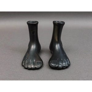 Può includere: Due vasi a forma di piede color bronzo scuro. Ogni vaso presenta un design dettagliato del piede con le dita e un'apertura cilindrica alla caviglia. I vasi sono esposti su una superficie grigia.