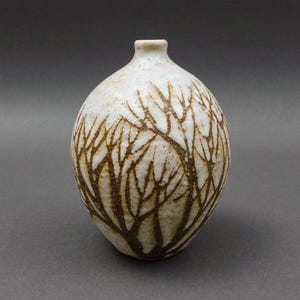 Puede incluir: Un jarrón de cerámica blanco con un diseño de árbol marrón. El jarrón tiene un cuerpo redondeado y un cuello estrecho.