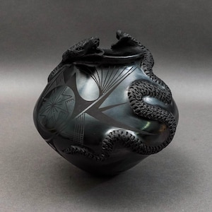 May include: A black ceramic pot with a detailed snake design. The pot features etched geometric patterns and a sculpted snake wrapping around the side and two snake heads on top. The pottery is a dark, matte black.