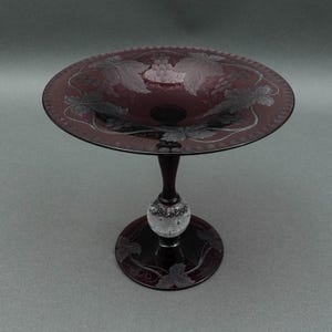May include: A burgundy-coloured, stemmed glass serving dish. The dish features a wide, shallow bowl with etched grape and leaf designs. The stem includes a clear, textured ball. The base also has etched leaf designs.