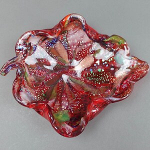 Puede incluir: Un cuenco de vidrio rojo con una forma ondulada e irregular. El cuenco tiene un acabado moteado e iridiscente con toques de oro, verde y blanco.