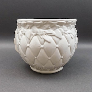 Puede incluir: Una maceta de cerámica blanca con una superficie detallada y texturizada. La maceta presenta un patrón de diamantes acolchados y un diseño de hojas decorativas alrededor del borde. La maceta tiene forma redondeada y una base pequeña y estable.