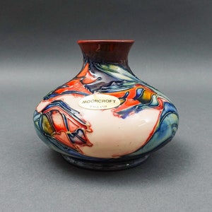 Puede incluir: Un pequeño jarrón redondo de cerámica de color crema con un borde rojo y un diseño abstracto azul y naranja. El jarrón tiene una etiqueta dorada que dice "Moorcroft England".