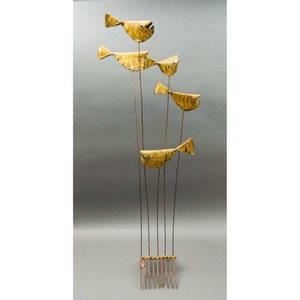 May include: A gold metal sculpture of five stylized fish suspended from thin metal rods. The fish are arranged in a vertical line, with the largest fish at the top and the smallest fish at the bottom. The sculpture is mounted on a base of metal rods.
