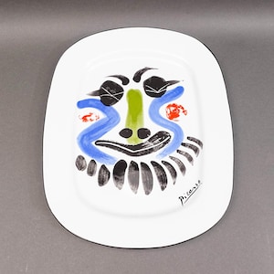 Op de afbeelding: Witte keramische schaal met een zwarte rand, met een kleurrijk abstract gezichtsontwerp. Het gezicht omvat zwarte ogen, een groene neus, blauwe lijnen en rode accenten. De schaal is gesigneerd "Picasso".