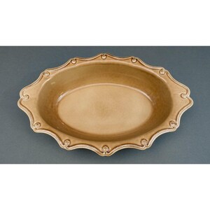 Peut inclure: Un plat de service ovale en céramique de couleur beige uni. Le plat présente un bord festonné avec une texture en relief rappelant une corde et de petits détails floraux circulaires espacés régulièrement sur tout le bord extérieur.